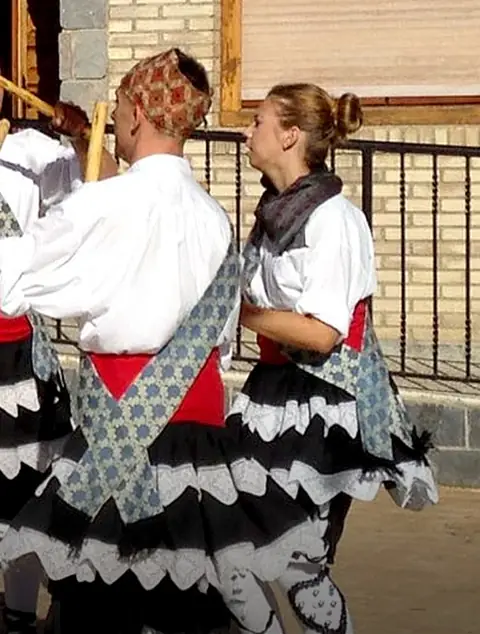 Danzantes con camisa blanca, fajín rojo y falda de volantes, bailando con palos en una calle de Maleján